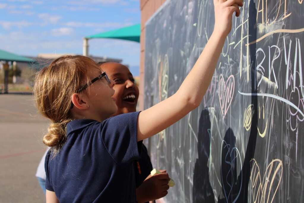 Two students use chalk on an outdoor chalkboard.